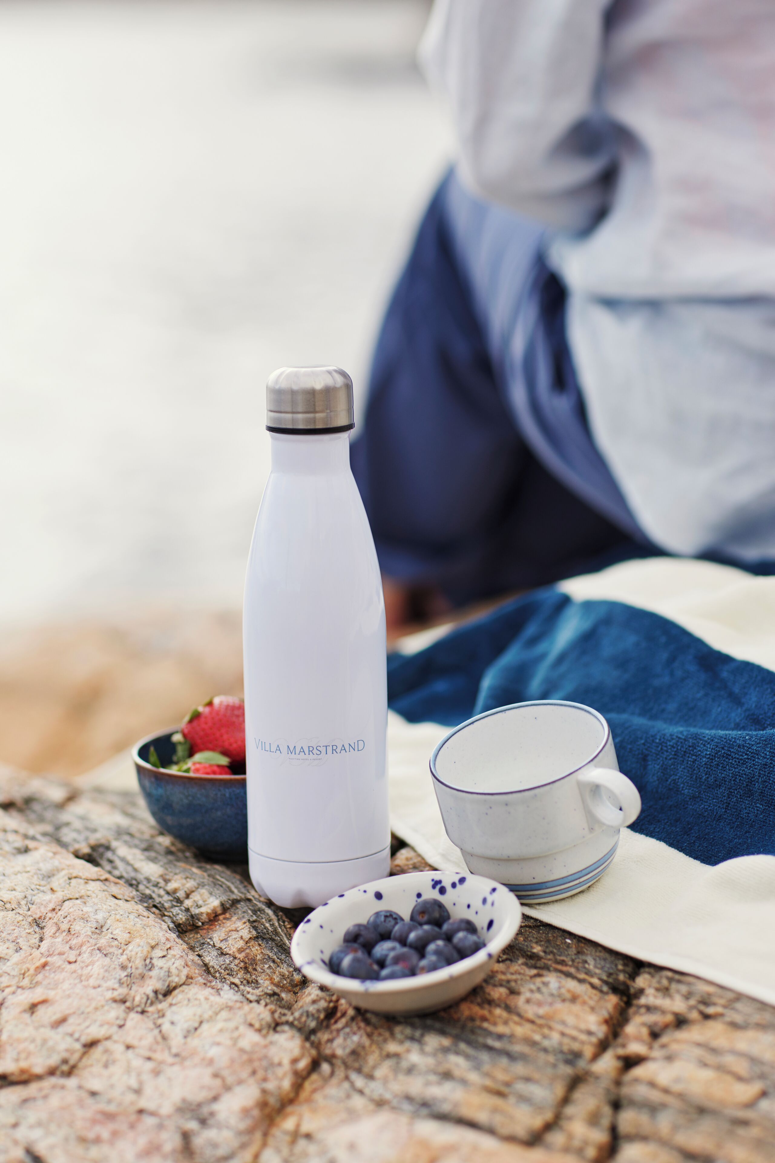 A white water bottle labeled "VILLA MARSTRAND" on a table with a bowl of strawberries, a bowl of blueberries, and a white mug.