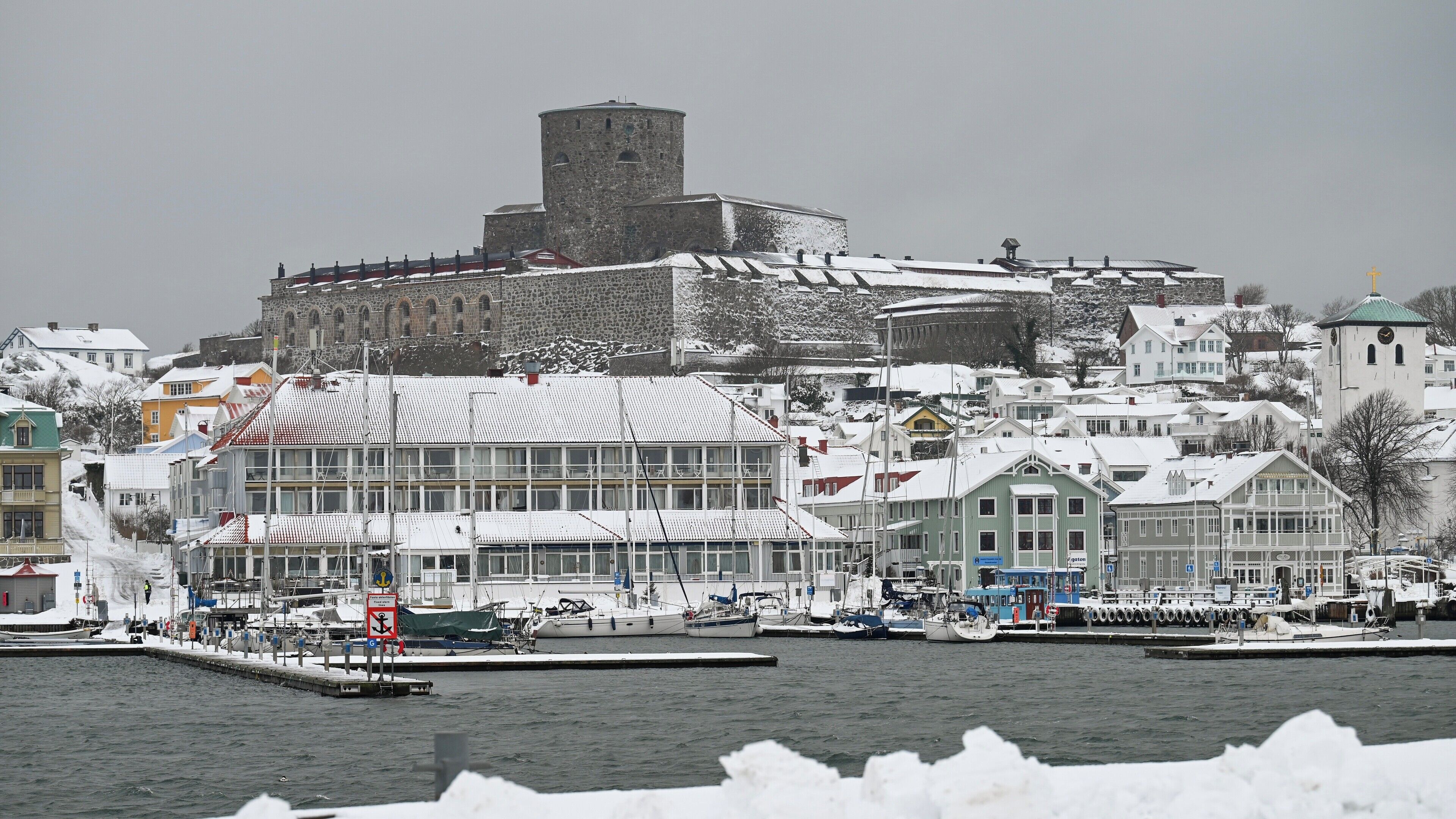A historic town with a prominent stone castle on a hill, modern buildings, and a marina with boats.