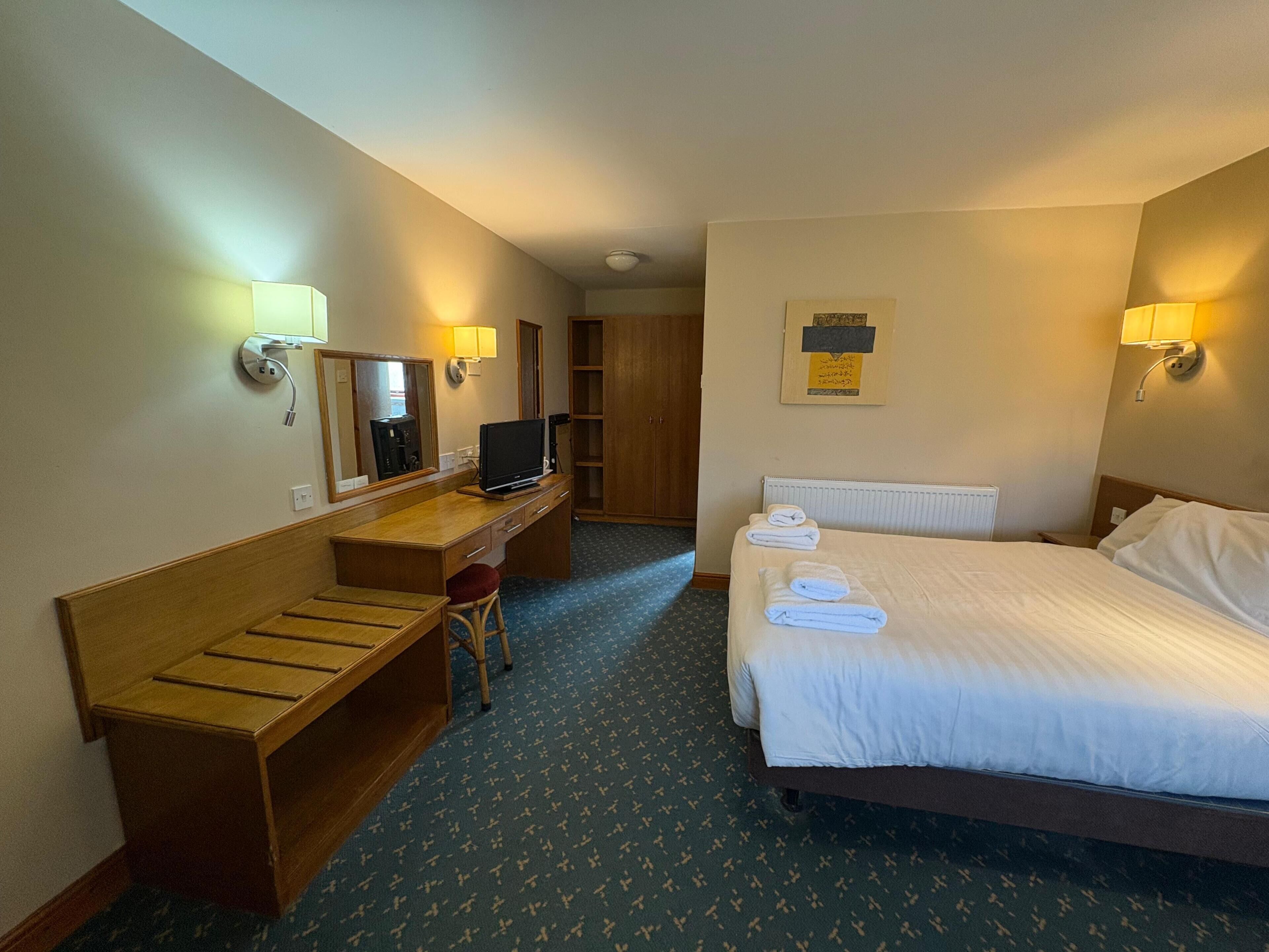 Ein Hotelzimmer mit einem Bett, einem Schreibtisch mit Stuhl, einem Fernseher und zwei Wandlampen.