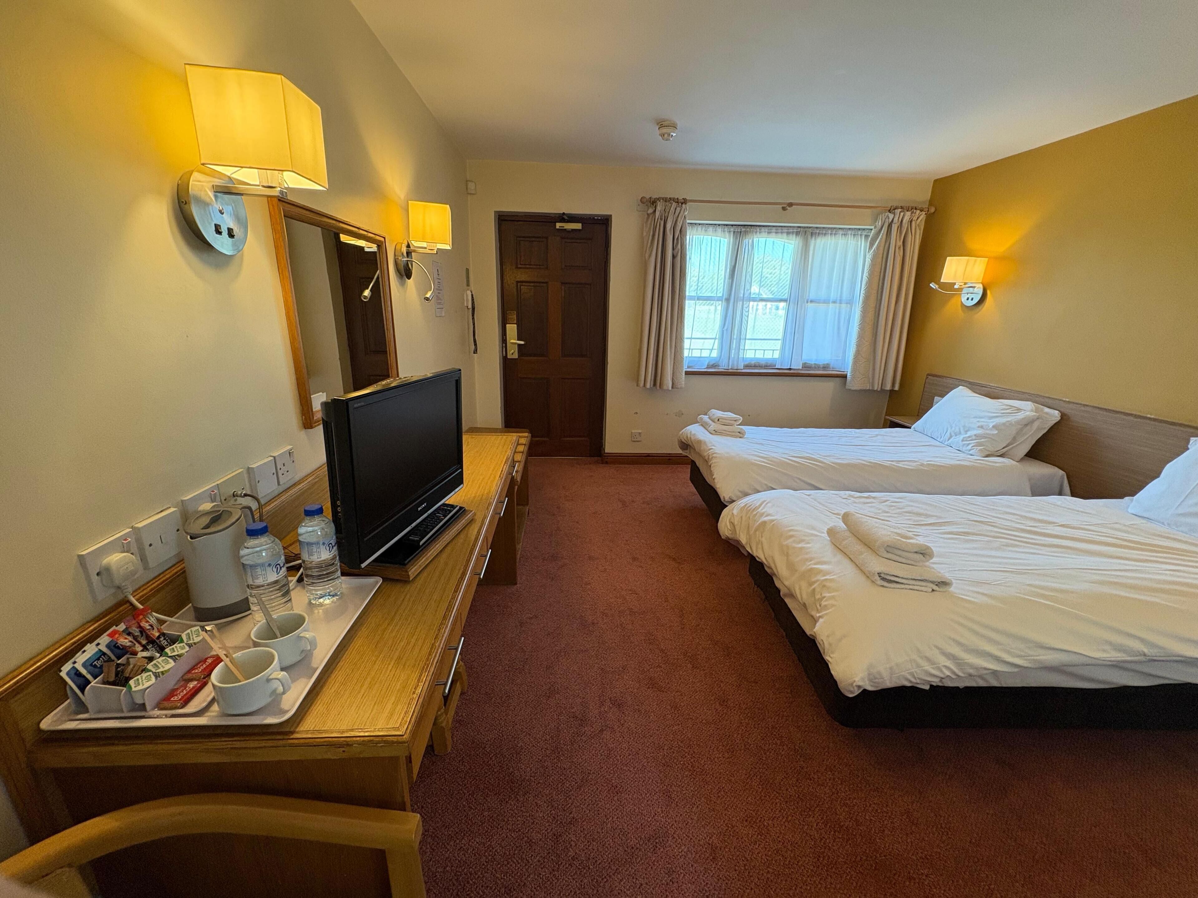 Een hotelkamer met twee bedden, een televisie op een houten tafeltje, een raam met gordijnen en een deur.