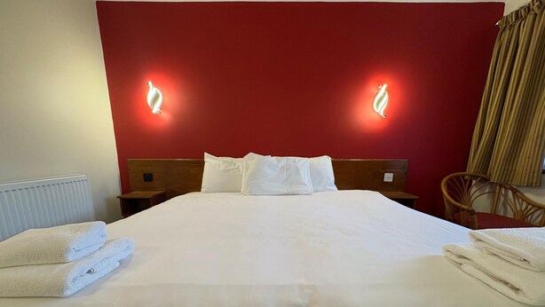A neatly made bed with white linens and pillows, two bedside tables, a wooden headboard, and two wall-mounted light fixtures. - Tollgate Hotel (Stoke-on-Trent)
