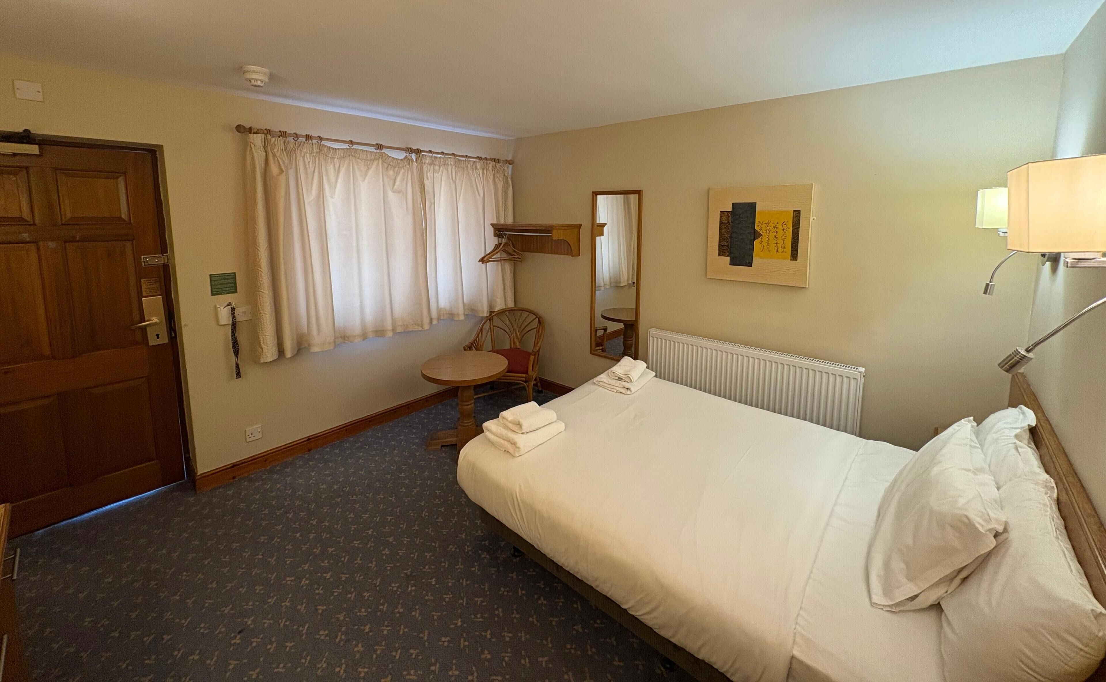 Ein Hotelzimmer mit Bett, Stuhl, Tisch, einem Fenster mit Vorhängen, einer Heizung und einer Wandlampe.