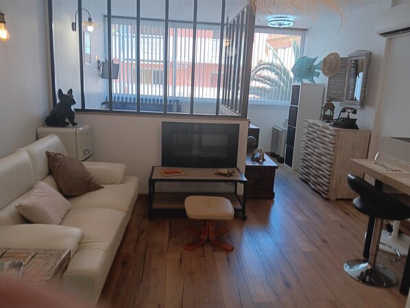 A living room with a sofa, a coffee table, a TV, and a small table with a chair. There is a window with blinds, a radiator, and a mirror on the wall.