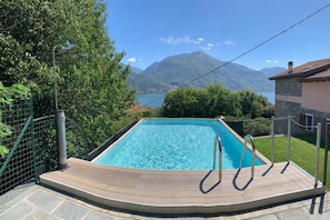 A rectangular swimming pool with a wooden deck and metal railings, surrounded by greenery and a mountainous backdrop. - Cremia Dreamhouse (Cremia)