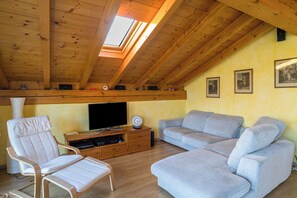 A loft-style room with a wooden ceiling, a skylight, a flat-screen TV, a grey sofa, a white chair, and a wooden entertainment unit.