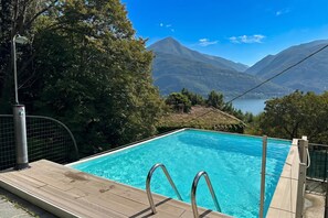 A swimming pool with a wooden deck, metal railings, and a view of mountains and a lake.