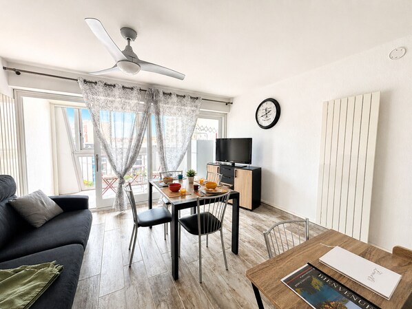 A modern living room with a dining area, a ceiling fan, a flat-screen TV, and a clock on the wall.