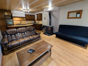 Una sala de estar acogedora con un sofá de cuero, un sofá oscuro, una mesa de centro de madera, una chimenea y una zona de cocina con armarios de madera y electrodomésticos.