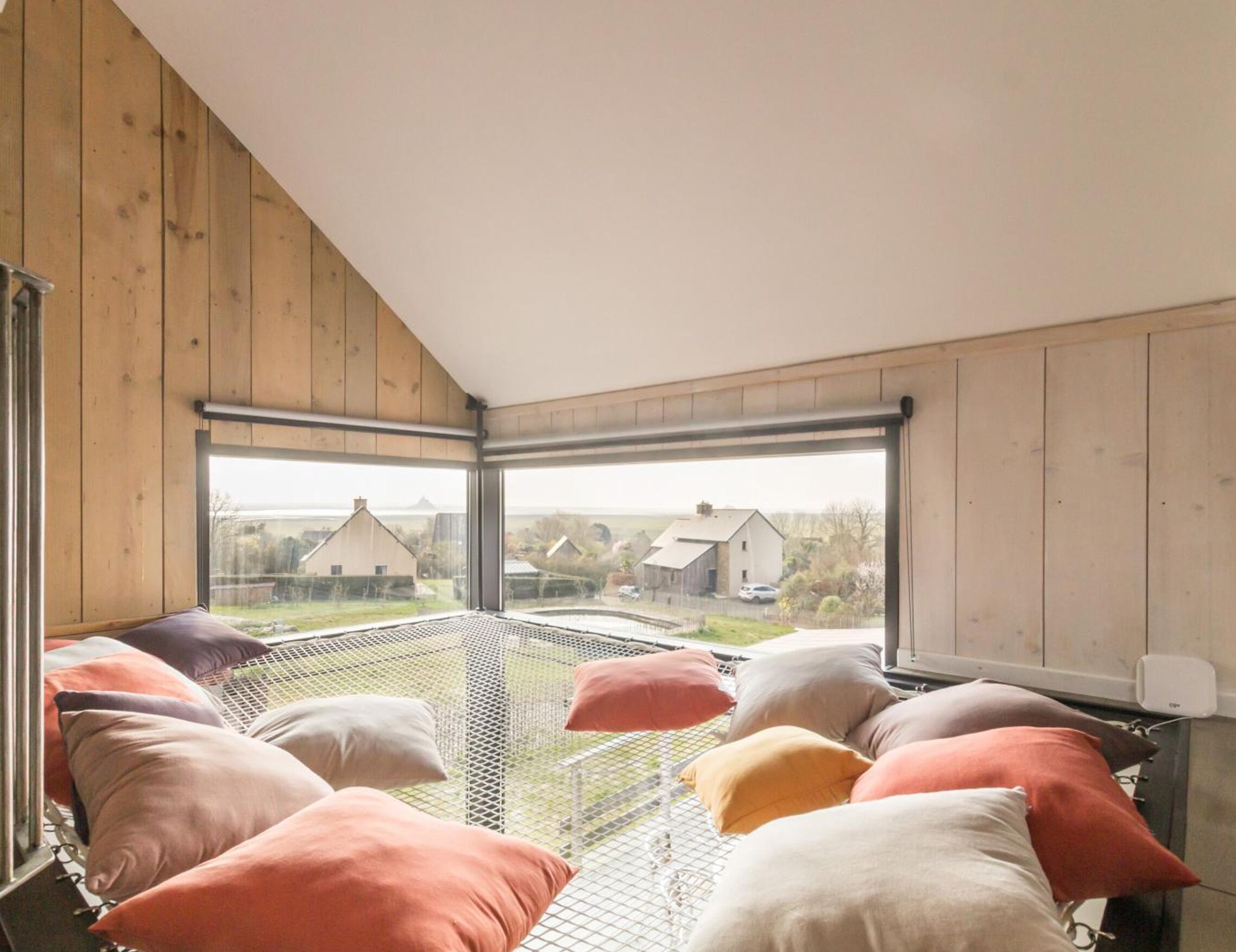 Une pièce avec un plafond haut, des murs en bois et une grande fenêtre donnant sur des maisons et une clôture.