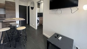 A modern kitchen with dark flooring, a dining area with stools, a flat-screen TV mounted on the wall, and a small table with a remote control.