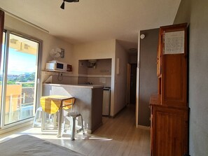 A compact living space with a kitchenette, a dining area with a table and chairs, a wooden door, and a view through a large window. - Studio with Terrace and Parking, Canet-en-Roussillon (Canet-en-Roussillon)