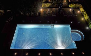 Una piscina rettangolare con una scaletta, circondata da sedie e illuminata da luci.