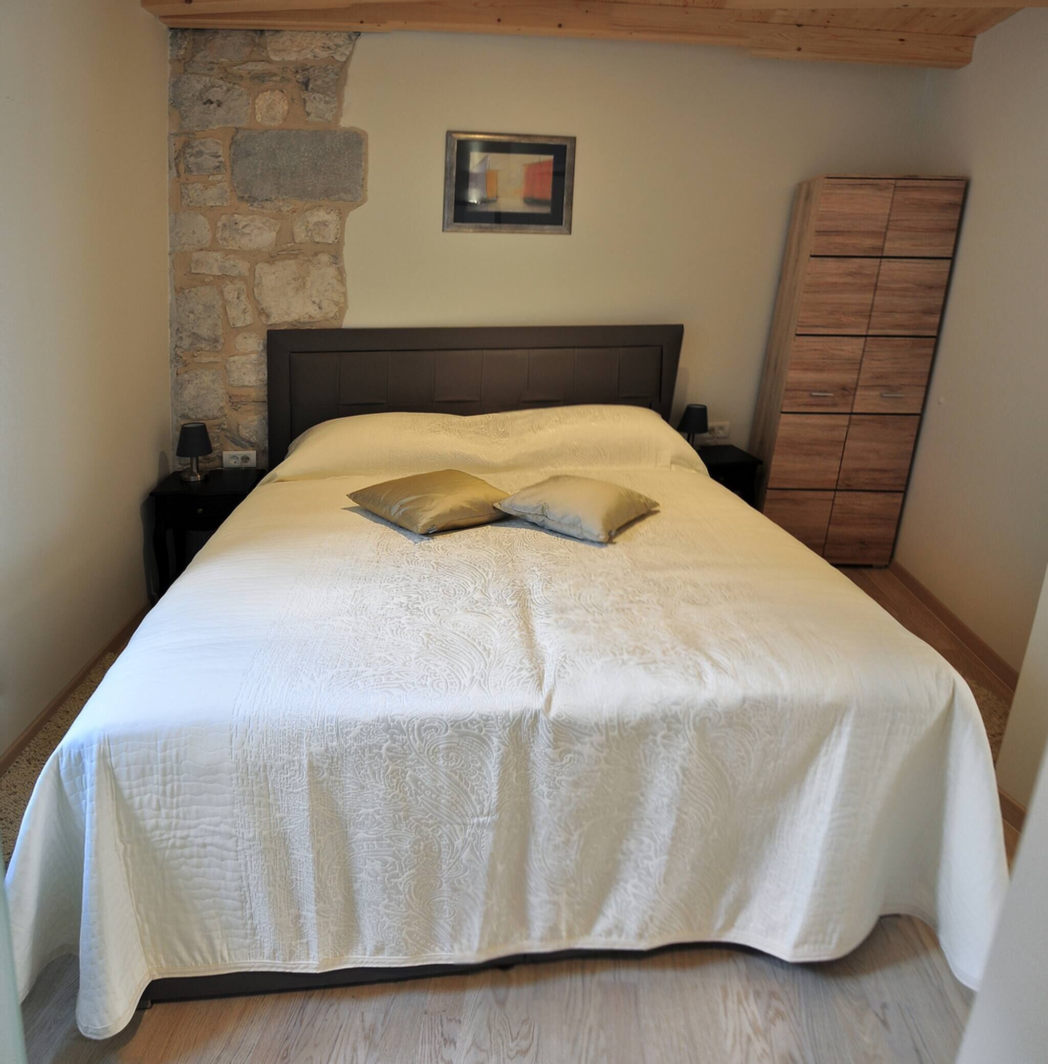 A neatly made bed with a white bedspread and two pillows, a wooden headboard, a stone wall, a framed picture above the bed, a nightstand with a lamp, and a wooden wardrobe.