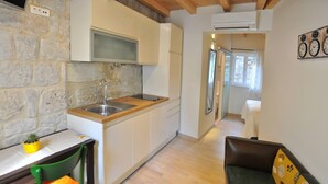 A compact kitchen with white cabinets, a wooden countertop, and a stone wall. There is a small table with a chair and a potted plant. A sofa is visible in the background. - Modern Studio in the City Center (Split)