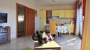 A living room with a television, a wooden cabinet, and a table with flowers. There is a kitchen in the background with yellow cabinets and a dining area. - Stunning View Suite With Balcony (Trogir)