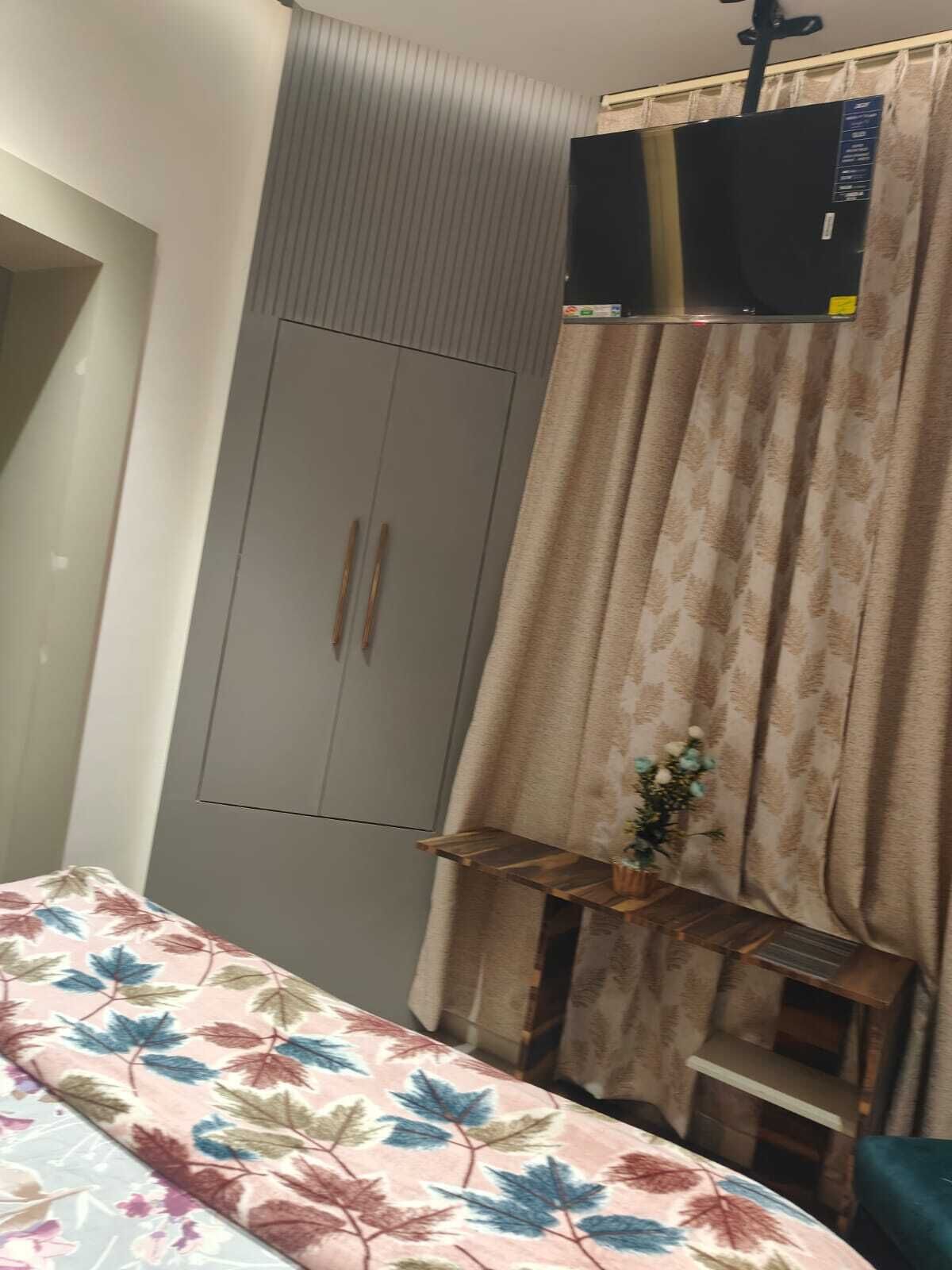 A bedroom with a grey wardrobe, a bed with a floral bedspread, a small shelf with a plant, and a window with curtains.