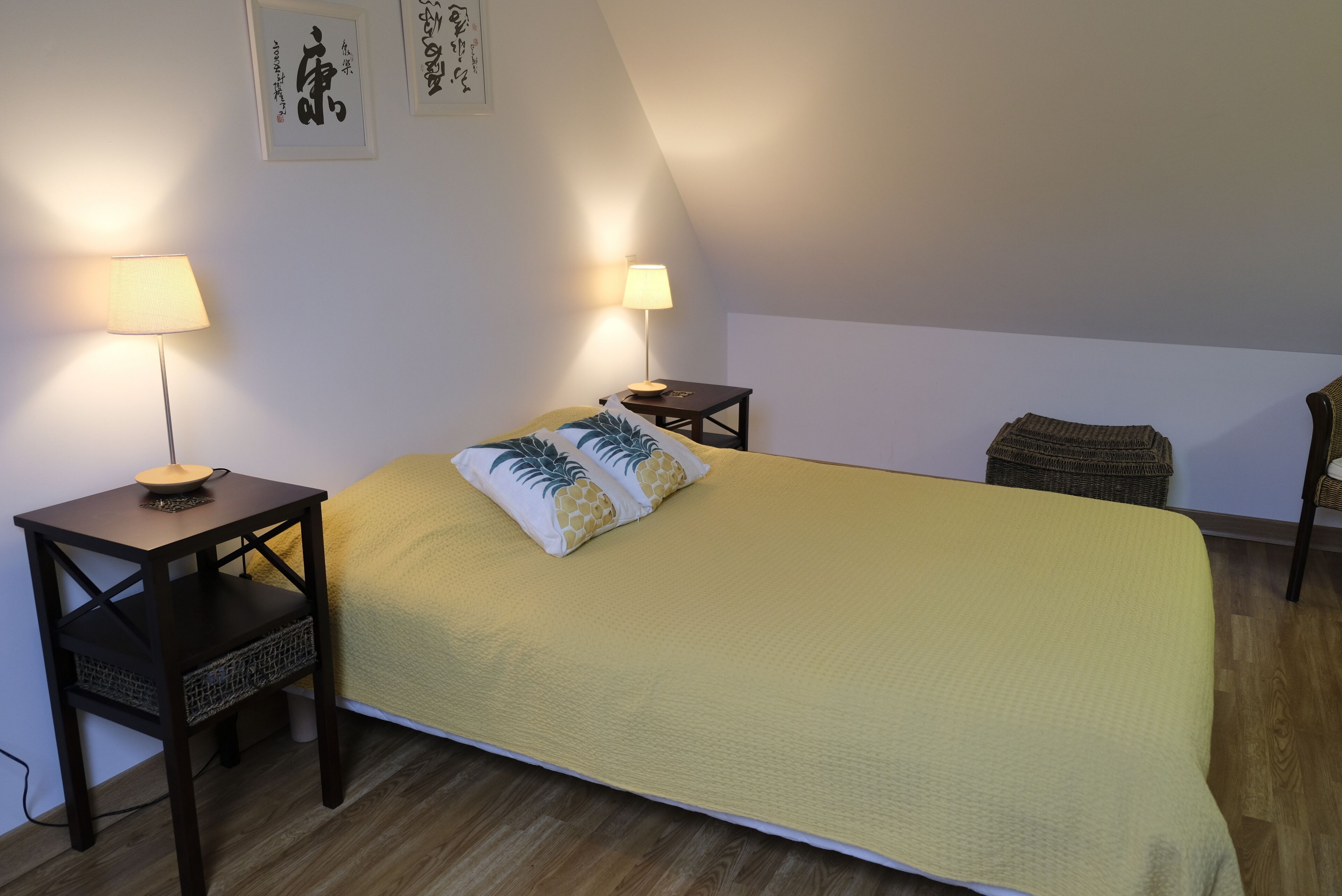 Ein Schlafzimmer mit einem großen Bett, zwei Nachttischen mit Lampen, einem Korbstuhl und Kunstwerken an den Wänden.