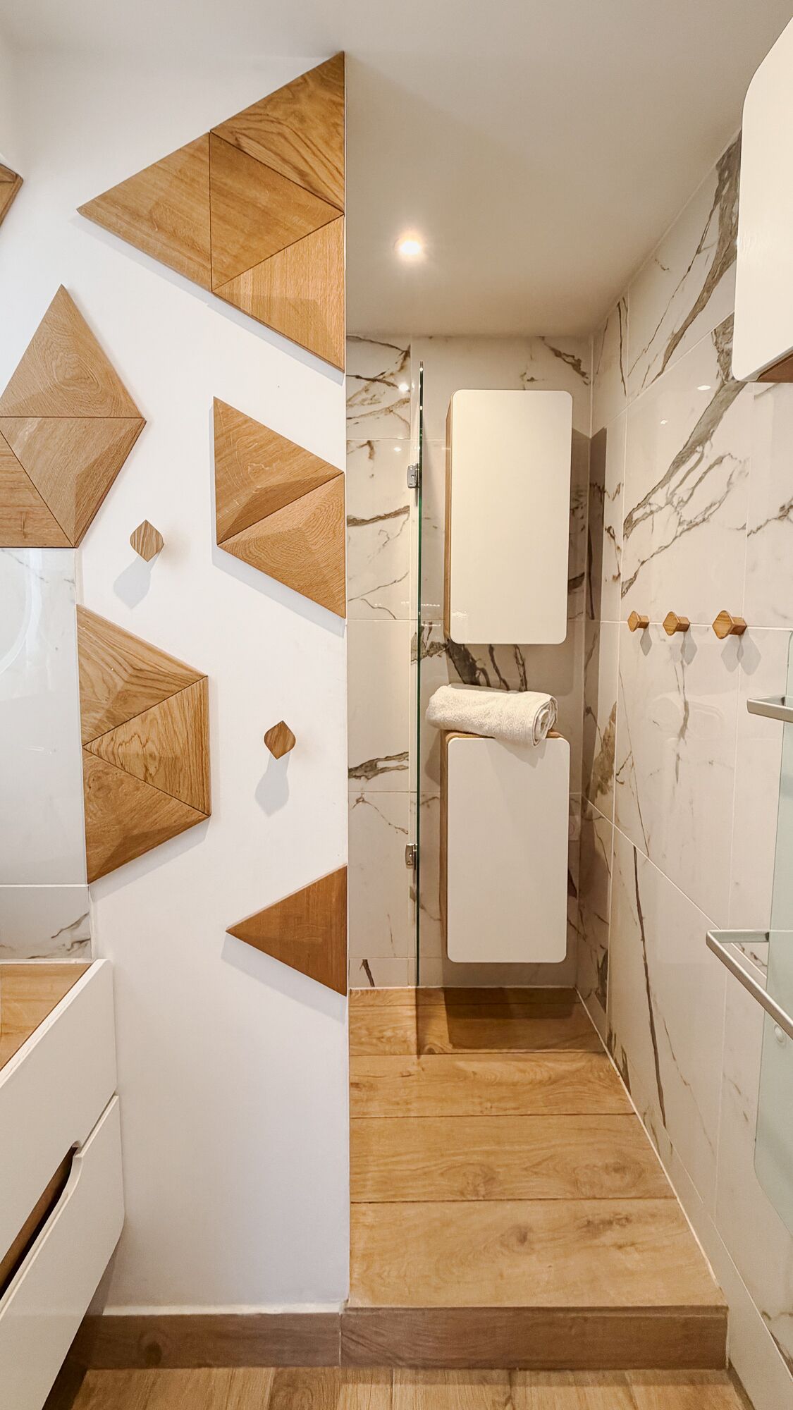 Un bagno moderno con un design geometrico a parete, una zona doccia rivestita in marmo e un pavimento in legno.