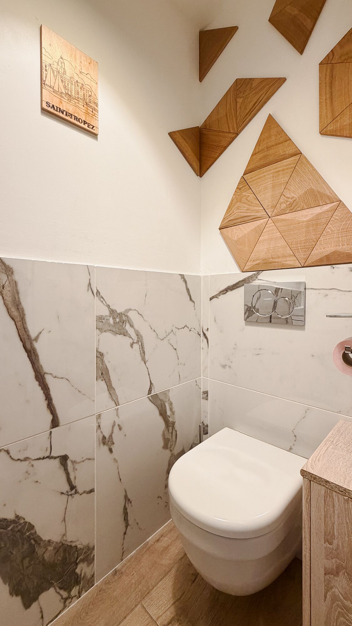 A bathroom with a toilet, marble walls, and geometric wooden wall decor.