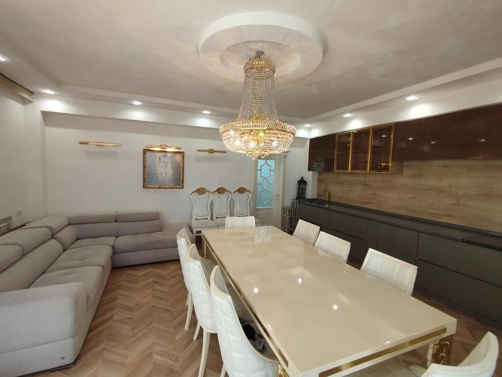 A modern dining room with a large marble table, white chairs, a chandelier, and a herringbone-patterned floor.