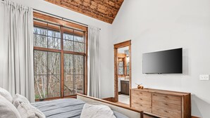 Un dormitorio con un ventanal grande, techo de madera, televisor y cómoda de madera.