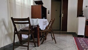 A room with a tiled floor, a wooden dining table with chairs, a small cabinet, and a door leading to another room.