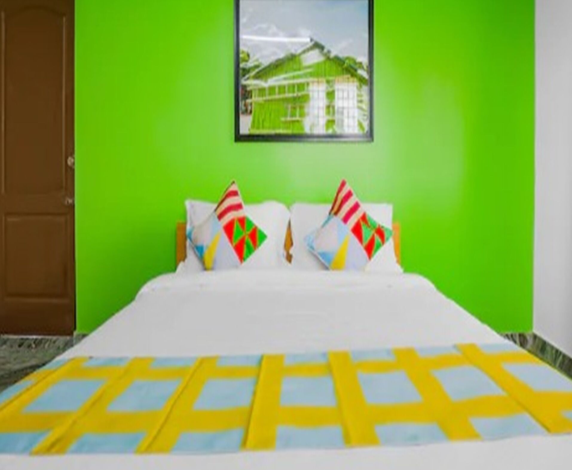 Una cama con una colcha a cuadros blancos y amarillos, dos almohadas con patrones geométricos coloridos y un cuadro de un edificio en una pared verde.