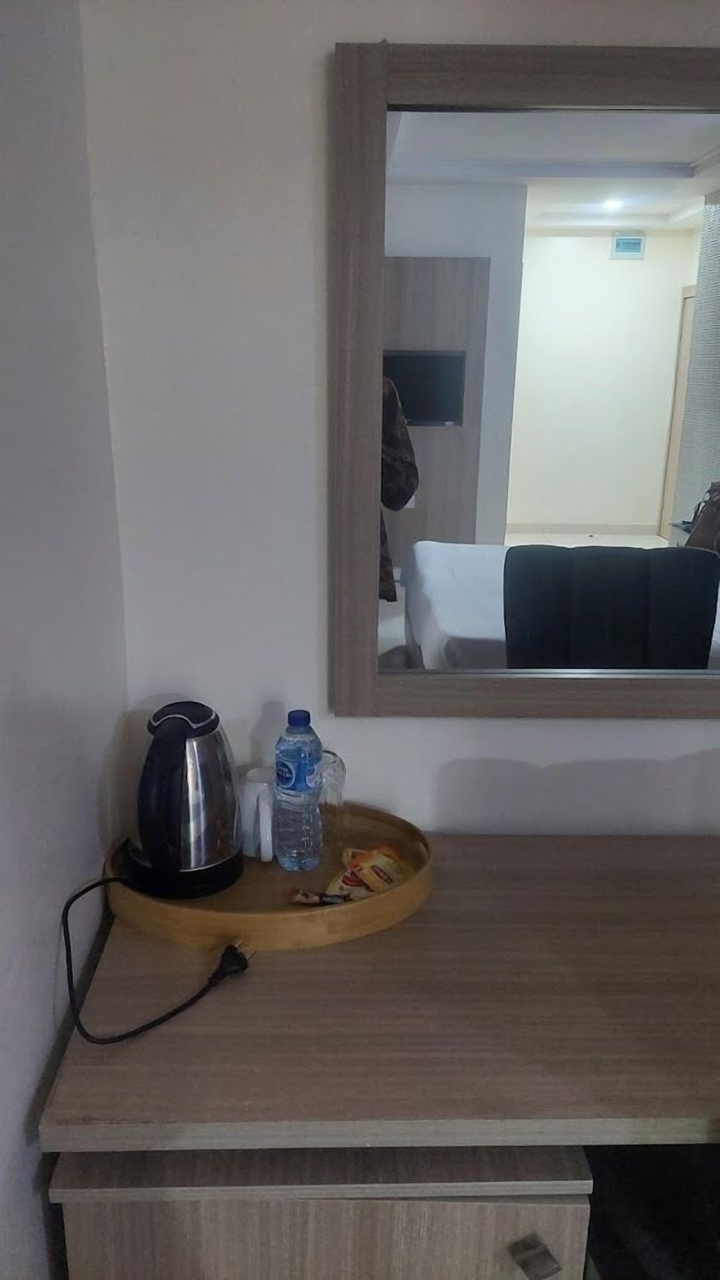 A wooden desk with a kettle, a water bottle, and a tray with snacks. A mirror reflects a room with a bed and a door.