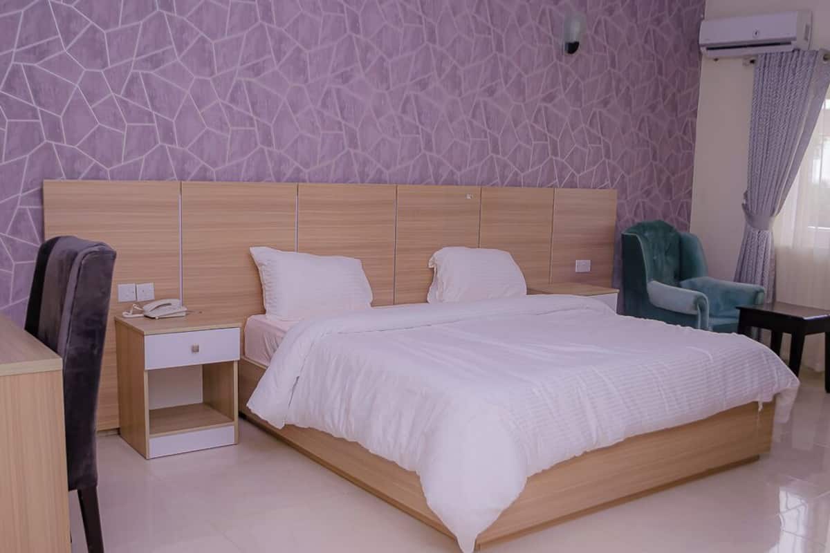 A modern bedroom with a large bed, a wooden headboard, a nightstand with a phone, a chair, and a small table.