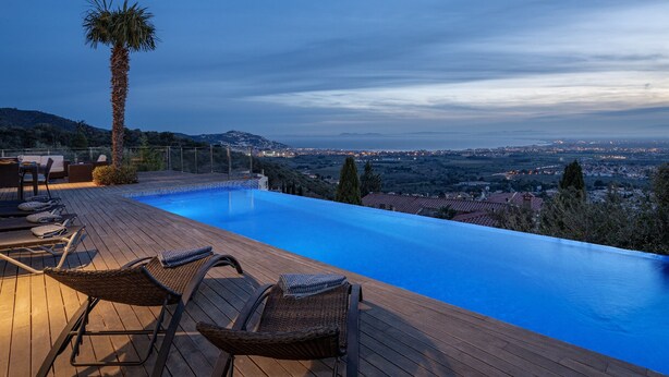 Una piscina a sfioro con zona prendisole in legno e una palma, affacciata su una città al crepuscolo.