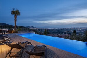 An infinity pool with wooden deck seating and a palm tree, overlooking a cityscape at dusk. - Casa Blanca - Exclusive villa with infinity pool and panoramic views (El Mas Fumats)