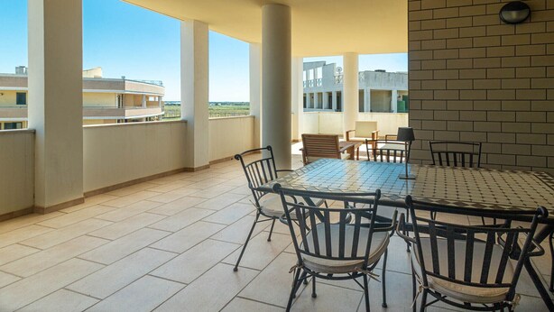 Une terrasse extérieure couverte, dotée d’une table ronde et de chaises, de grandes colonnes et offrant une vue sur les bâtiments en arrière-plan.