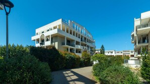 Un immeuble moderne à plusieurs étages, doté de balcons, entouré de verdure et surplombant un ciel bleu azur.