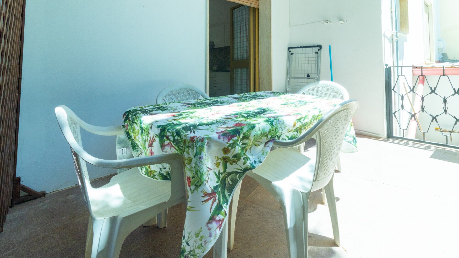 A dining area with a table covered in a floral tablecloth, surrounded by white chairs.