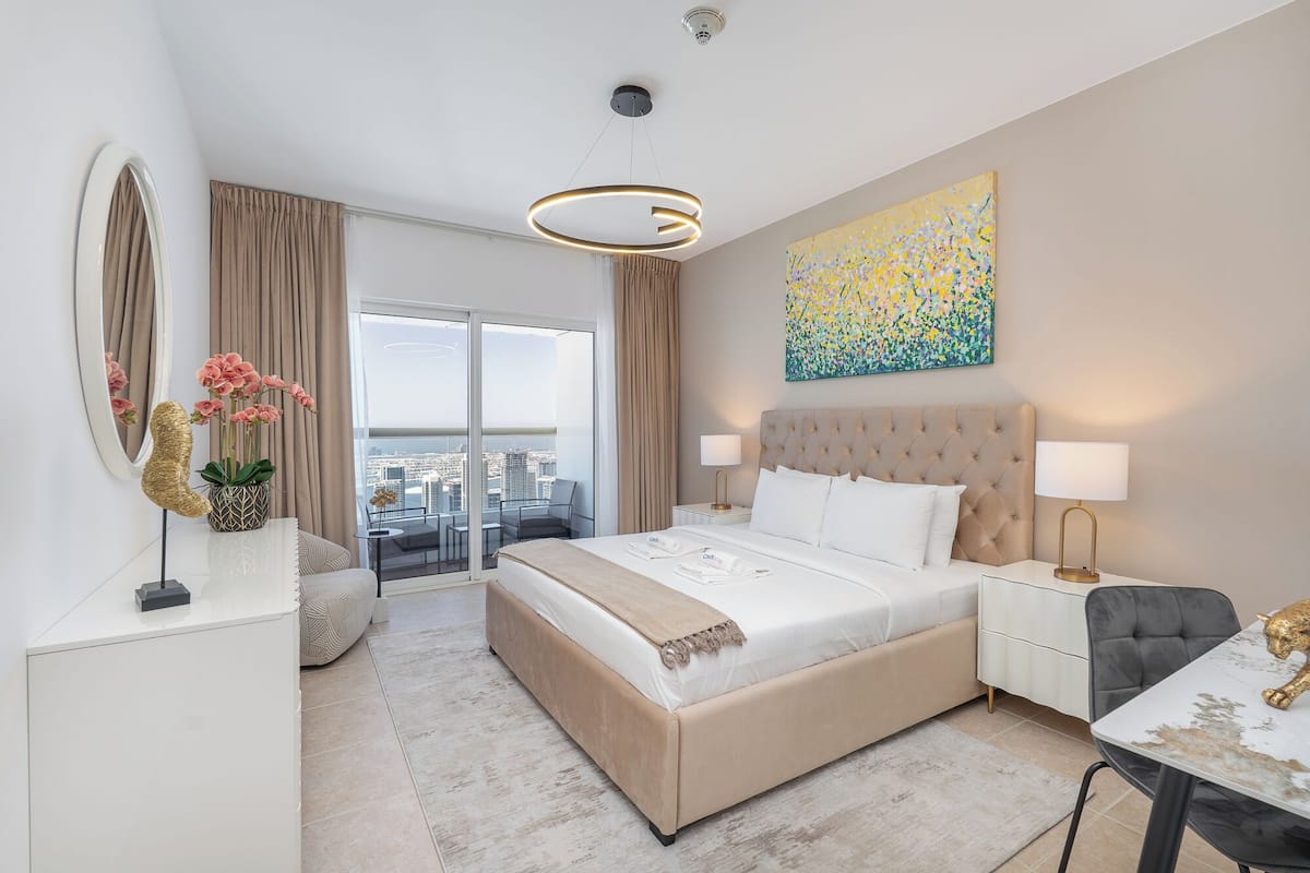 Una habitación de hotel moderna con una cama grande, dos mesitas de noche, un escritorio, una silla, un ventanal con vista a la ciudad y un cuadro abstracto colorido en la pared.