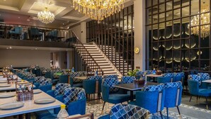 A spacious dining area with multiple tables and blue upholstered chairs, a grand chandelier, and a staircase leading to an upper level. - Saltstayz Autograph Connaught Place (New Delhi)