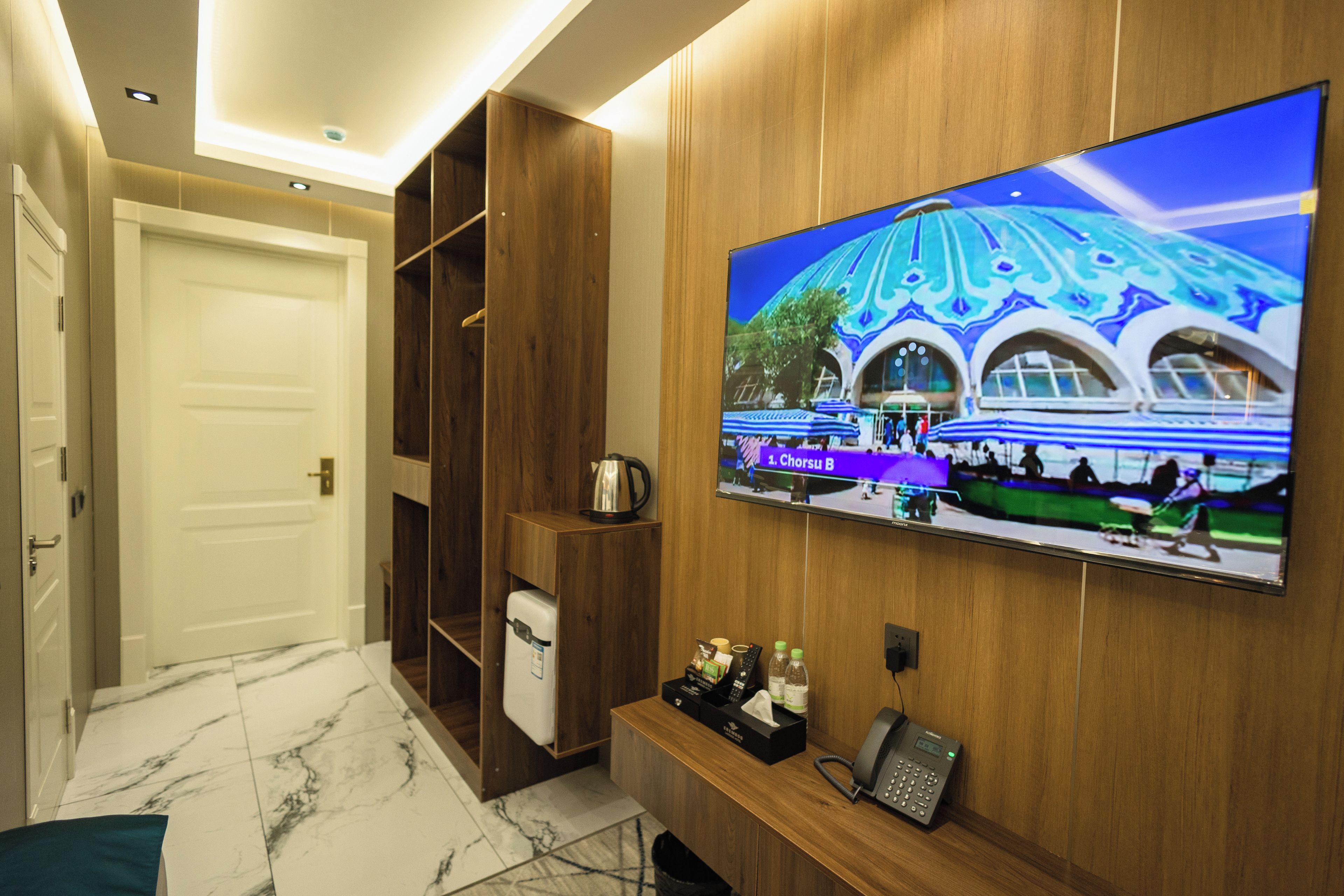 A hotel room with a large flat-screen TV mounted on the wall, a wooden cabinet, a small refrigerator, and a telephone.
