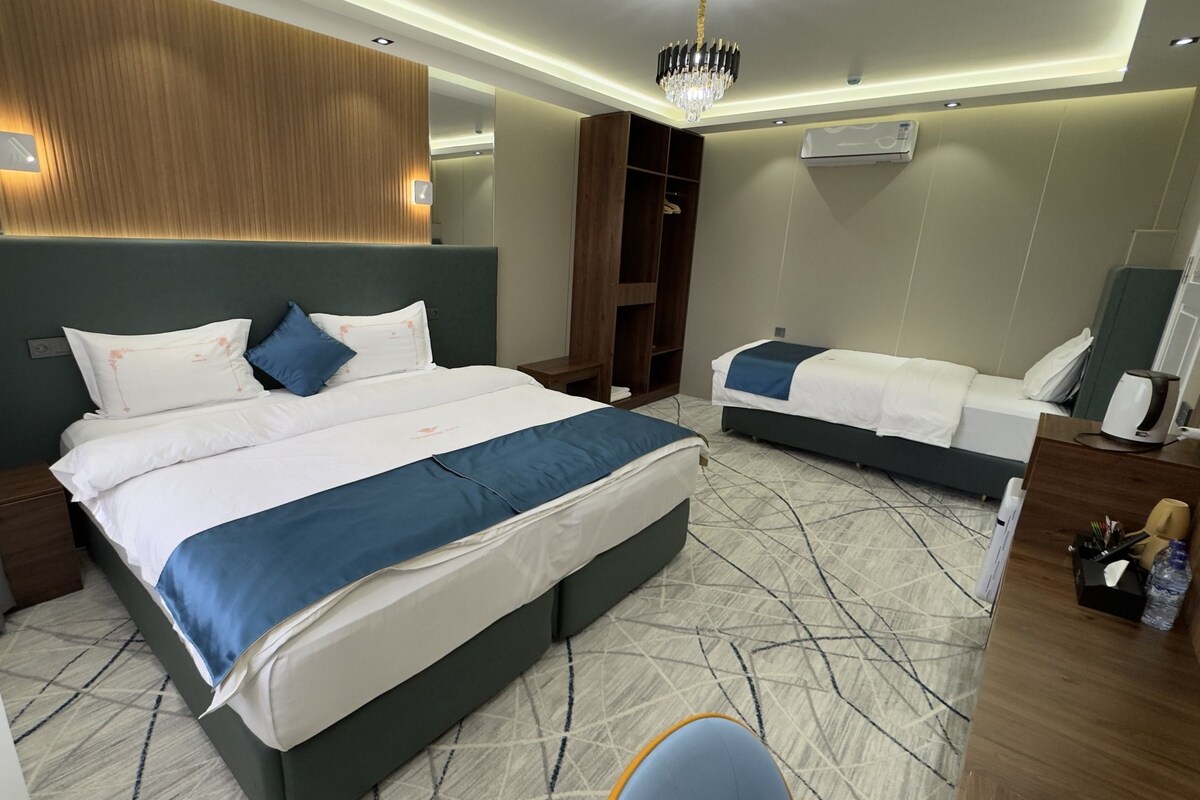 A hotel room with a large bed, a smaller bed, a wardrobe, a desk with a chair, and a chandelier.