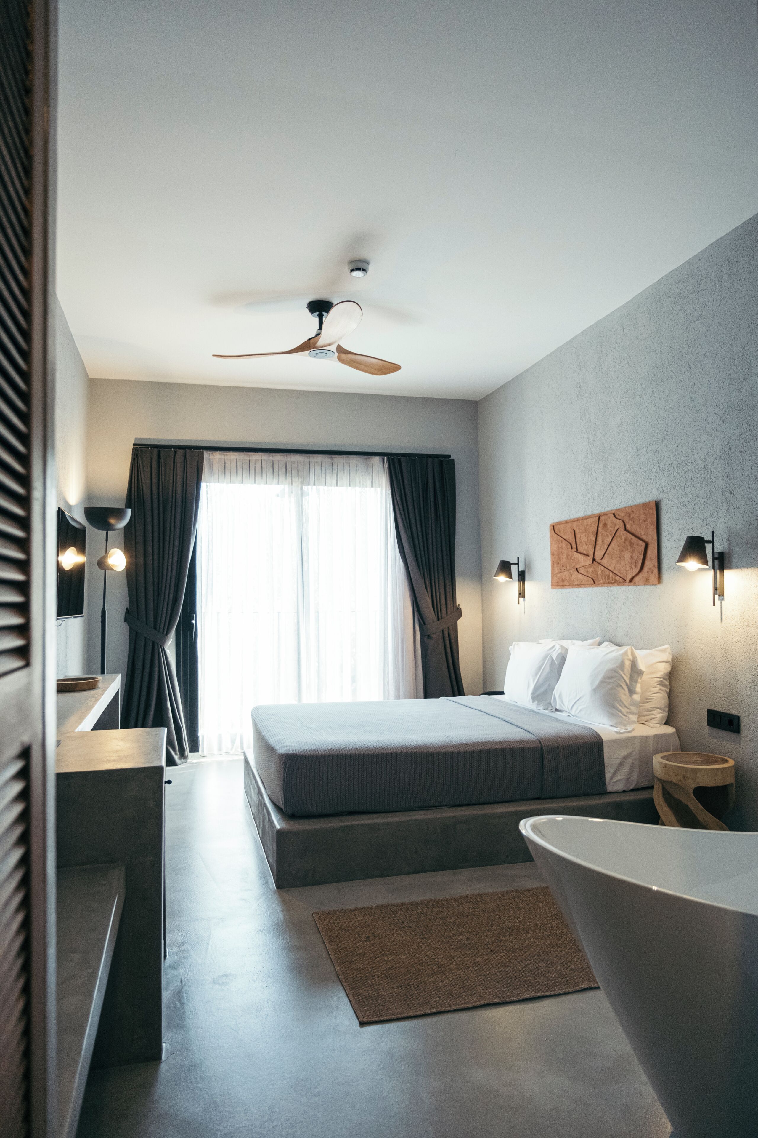 Quarto de hotel moderno com cama, banheira, escrivaninha e uma janela grande com cortinas.