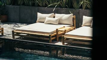 Dos camas de madera con ropa blanca, una jardinera de concreto con plantas verdes grandes y una piscina cristalina con barandilla de metal.