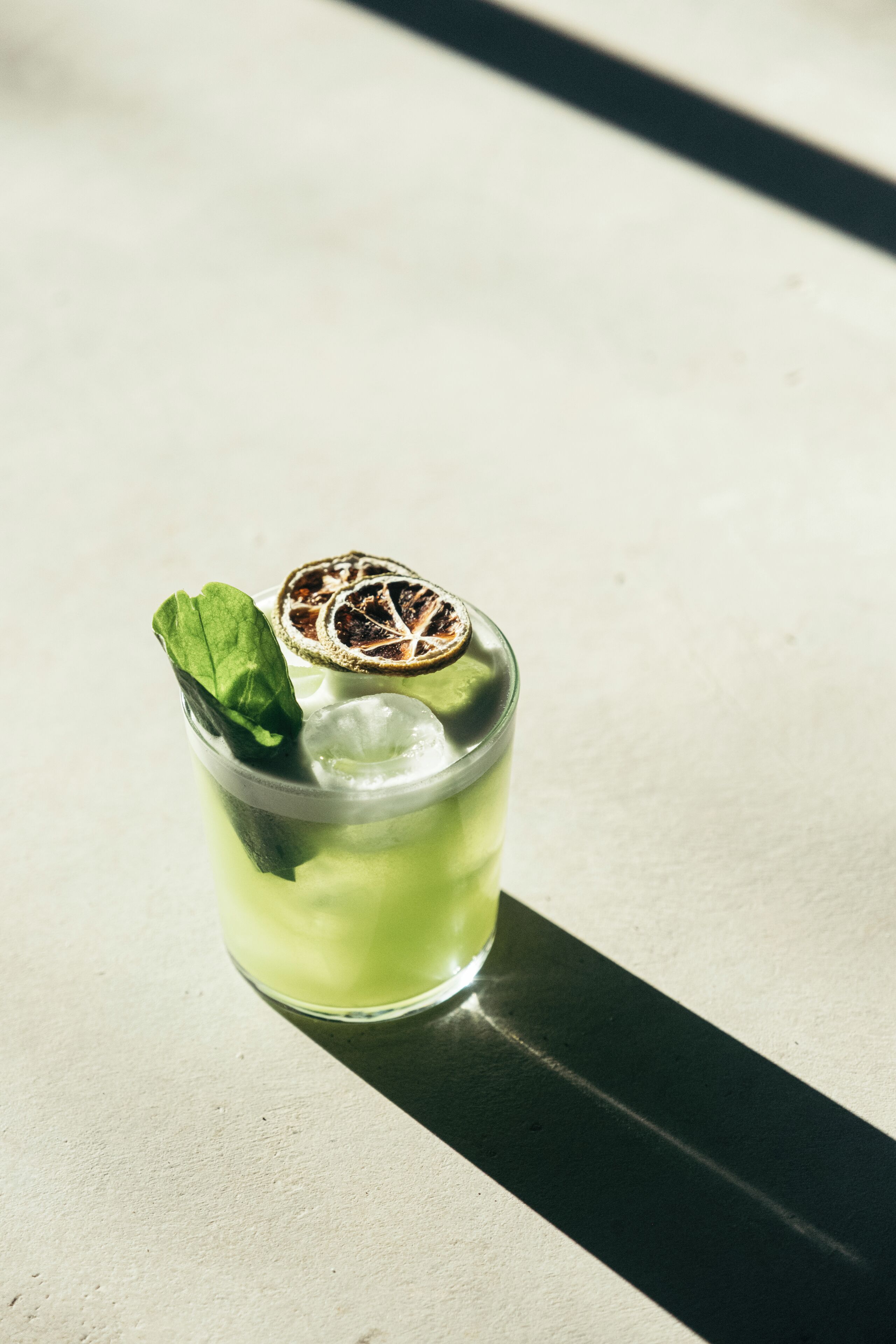 A clear glass with a green beverage, ice cubes, a mint leaf, and a dried citrus slice garnish on a light-colored surface.