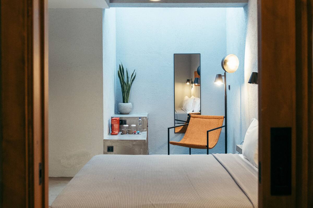 A bedroom with a bed, a chair, a small table with a plant, a mirror, and a lamp.