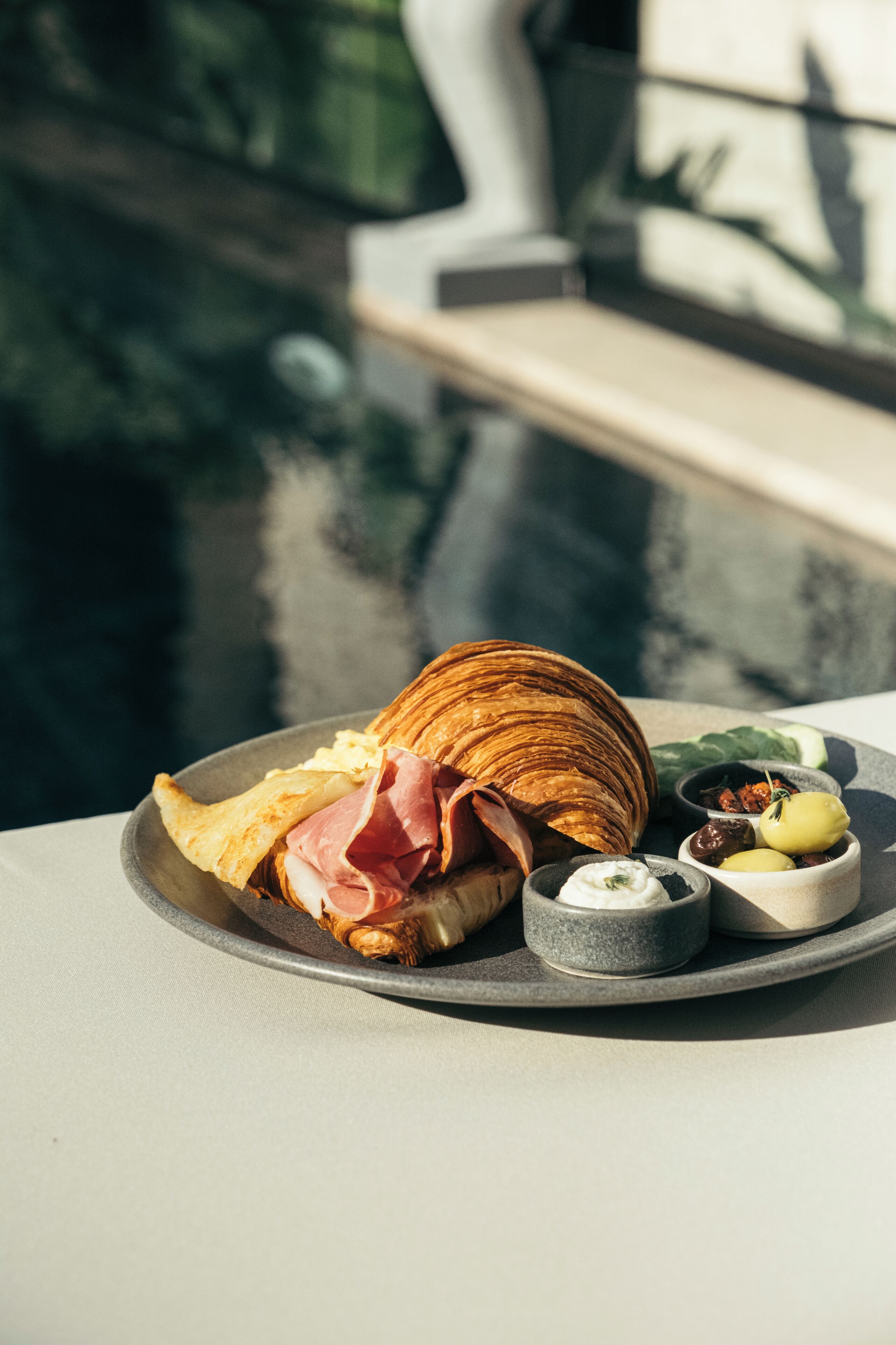 A breakfast plate with croissant, ham, and olives on a table by a pool.