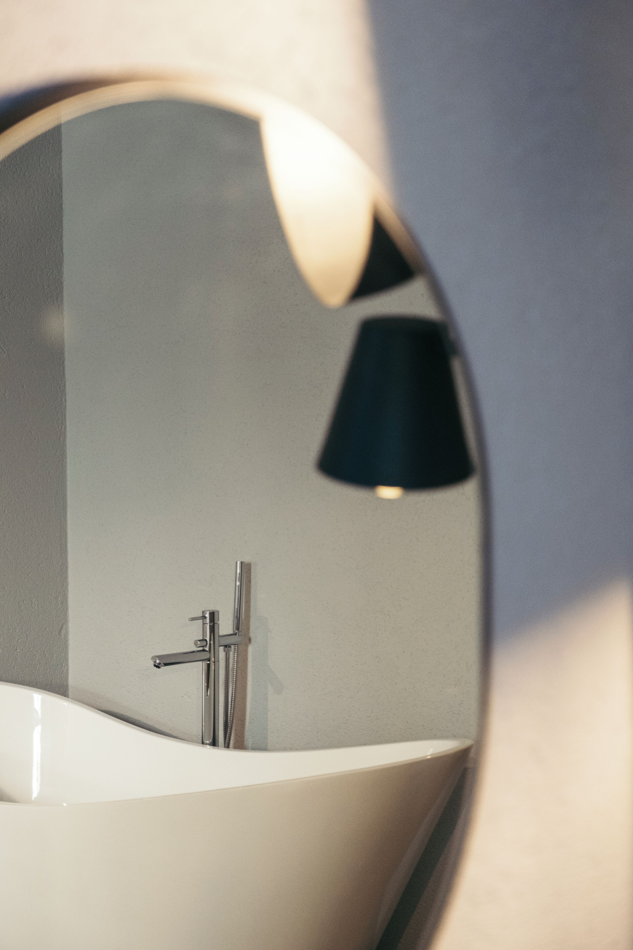 A bathroom with a round mirror, a white bathtub, and a black lamp.