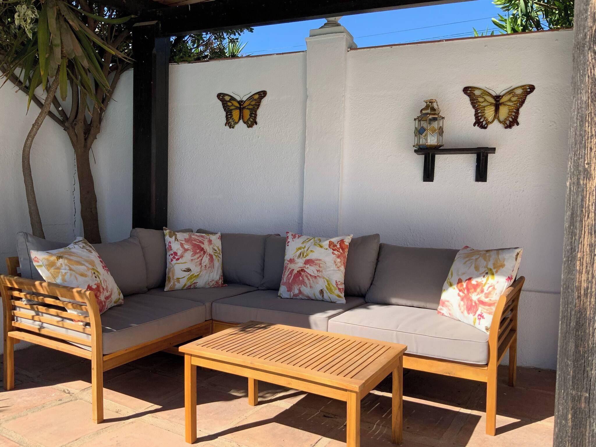 An outdoor seating area with a wooden table and two chairs, a sofa with floral cushions, and two butterfly wall art pieces.