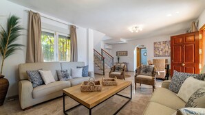 Un salon spacieux avec un canapé beige, une table basse en bois, des fauteuils en osier et une grande fenêtre agrémentée de rideaux.