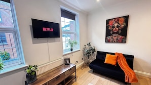 Un soggiorno con un divano nero, un mobile TV in legno, una TV a parete e una finestra vista un edificio in mattoni.