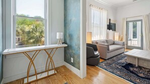 Une pièce avec une grande fenêtre, un bureau, un canapé gris et un tapis à motifs.