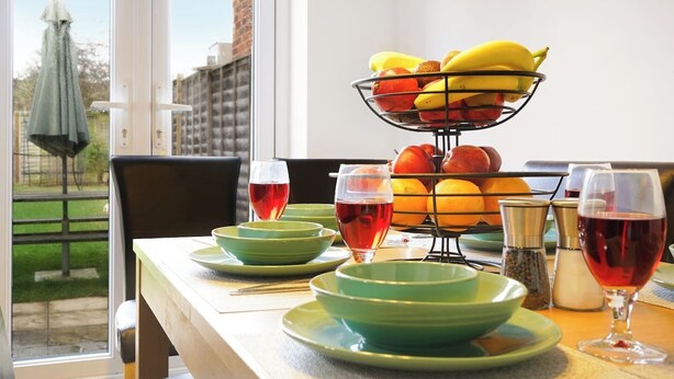 Una mesa de comedor con platos verdes, tazones y copas de vino tinto, una cesta de frutas con plátanos y manzanas, y vistas a un edificio y vegetación al exterior.