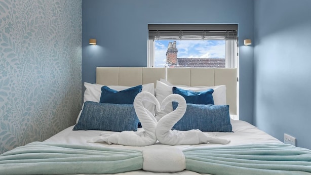 Ein Schlafzimmer mit einem Bett, blauen Kissen, einem weißen Kopfteil und einem Fenster mit Blick auf ein Gebäude.